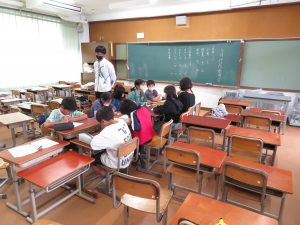 教室の中央の机を向い合わせに設置して10名の児童が座っており、先生が見守る中委員会活動が行われている様子の写真