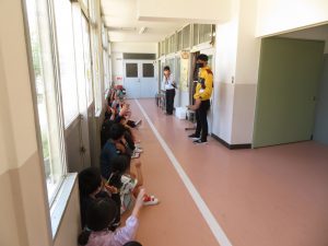 廊下の壁に沿って座っている子どもたちが先生の話を聞き右手を挙げて返事をしている様子を写した写真