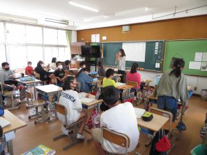 教室で教壇に立って授業をしている先生、自席についている生徒の中の一人が席を立ち発表しており、他の生徒が発表している生徒に注目している様子の写真