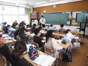 板書された黒板、子どもの様子を見ている白いワイシャツ姿の先生、机の上に教科書とノートを広げ自席に座って授業を受けている子どもたちの様子を写した写真