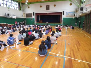体育館にたくさんの児童が集まり、整列して座っており、前方中央に一人の先生が立っている全体集会の様子の写真