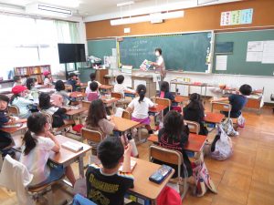 教室の黒板の前で授業を進めている先生と自席に座り先生の話を聞いている子どもたちが写る授業の様子の写真