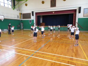 体育館で、先生と体操服を着た子どもたちが間隔をあけて広がっており、左手を頭にのせて首を傾け体操を行っている様子の写真