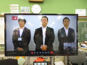 モニターに3人のスーツ姿の関係者の男性が写っているリモートでの総会の様子の写真