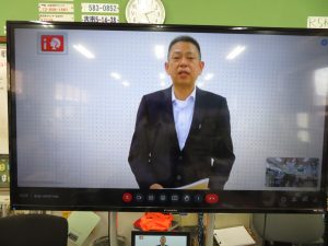 資料を手に持ち話をするスーツ姿の男性がモニターに映っている総会の様子の写真