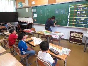 机の上に教科書やドリルがだされた自席に座っている3人の児童と黒板に板書している先生が写っている授業風景を写した写真