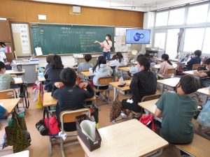 板書された黒板を両手で示しながら話をしている先生と、自席について先生の話を真剣に聞いている子どもたちの授業風景を後方から写した写真