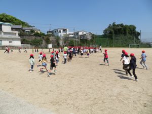運動場の一角に赤い帽子をかぶった児童が整列しており、次々と他の児童たちが運動場に走って集まっている避難訓練の様子の写真