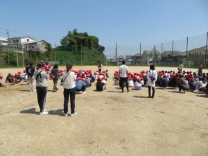 赤い帽子をかぶった児童たちが整列して座っており、一人の先生が子どもたちの前に立って話をしており、他の先生方も児童の後ろに立って話を聞いている避難訓練の様子を後方から写した写真