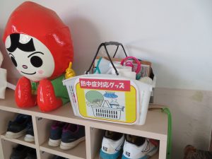 靴箱の上に「熱中症対応グッズ」と書かれ、必要な道具が描かれたイラストが貼ってある道具の入ったかごが「人KENまもる君」のマスコットの横に置かれている写真