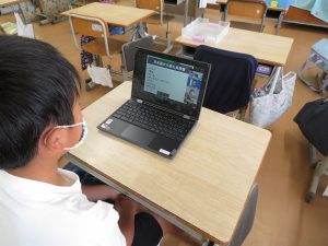 机の前に座っている男子児童がタブレットを広げており、タブレット画面にオンライン国際クラブOSAKAの会議の映像が映し出され、ウェブ会議を行っている様子の写真