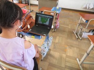 机の間に座っている女子児童がタブレットを広げており、タブレットの画面には地図の映像が写し出されウェブ会議を行っている様子の写真