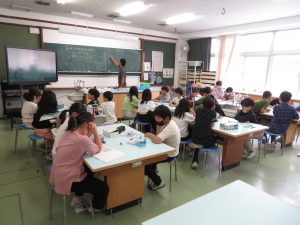 理科専科の男性教師が黒板に板書しており、4人掛けの机6台に子どもたちが座って授業を受けている様子の写真