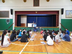 体育館で4年生の子どもたちが整列して座っており、舞台前で、警察館の男性が自転車を持って話をしている交通安全教室の様子の写真