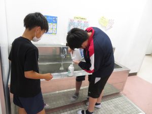 一人の男子児童が側で見守る中、右側の男子児童が手洗い場で、手洗い石鹼の容器にせっけん液の補充をしている様子を写した写真