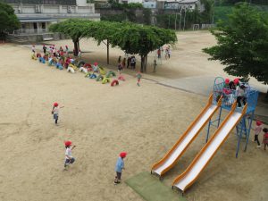 校庭に設置されている滑り台で遊んでいる複数の児童や藤棚近くで遊んでいる複数の児童など思い思いに遊んでいる児童たちの様子を写した写真