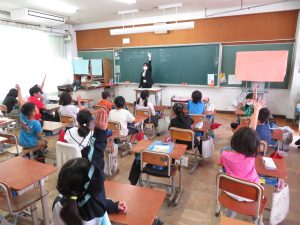 教室で黒色のスーツを着た実習生が教壇に立ち子どもたちと向かい合って左手を真っ直ぐ上に挙げており、自席に座っている子どもたちも元気よく手を真っ直ぐに挙げている実習生研究授業の様子の写真