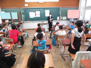 教室でで黒色のスーツを着た実習生が授業を行っており、自席に座っている子ども達の中の一人の女子児童が、自席に立ち発表をしている様子を後方から写した写真