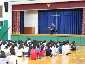 体育館の舞台には日本胡弓の演奏者の男性が椅子に座って楽器を持って話をしており、児童の皆さんが座って話を聞いている講演会の様子の写真