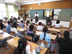 教室の黒板の前に映像が映し出されたモニターが設置され、男性教師が授業を行っており、自席に座っている子ども達が先生の話を聞いている授業風景の写真