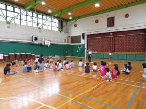 体育館に子ども達が整列して座っており、前に一人の先生が立って話をしている支援学級公開授業の様子の写真