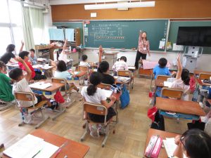 教室で黒板の前に女性教師が立って授業を行っており、自席に座っている子ども達が手を真っ直ぐに挙げている授業風景の様子を後方より写した写真