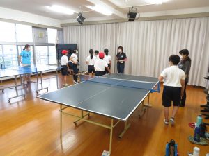 室内に2台の卓球台がおかれ、体操服を着た子どもたちが先生の周りに集まり話を聞いている卓球クラブの様子の写真