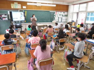 教室で子どもたちは自席についており、エプロンを付けた歯科衛生士さんが子どもたちの前に立って、身振り手振りで話をしており、子ども達も熱心に聞いている歯のみがき方教室の様子を後方より写した写真