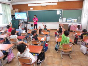 教室で女性教師が黒板の前に立ち、自席について授業を受けている子ども達が元気よく右手や左手を挙げている授業風景の様子を後方から写した写真