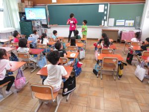 教室で黒板の前に、女性教師と男子児童が立っており、クラスメートの子どもたちは自席について興味深く2人の話を聞いている授業の様子を後方から写した写真