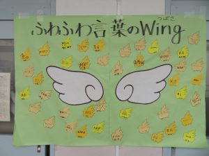 若草色の大きな模造紙に「ふわふわ言葉のWing（つばさ）」と書かれ中央に白い翼の絵が描かれ、まわりに色々な言葉の書かれたカードが張られ掲示されている写真