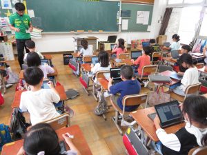 教室の自席に子どもたちが座っており、机にはそれぞれタブレットが置かれ、男性教師が黒板の前に立って授業が行われている様子の写真