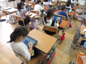 教室の後ろの席に座っている女子児童が、タブレットにタッチペンを使って文字を書き込んでいる様子を後方から写した写真
