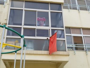 校舎の窓に赤帽をかぶった校長先生のパネルが出され、赤色の旗が窓から出されている様子の写真