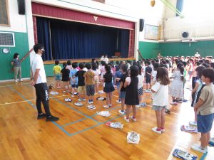 体育館で、子どもたちが整列して並んでおり、舞台前に立つ先生の話を聞いている全体集会の様子の写真