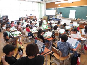 教室の自席に着いた子どもたちが、机の上に鍵盤ハーモニカを置き、授業を受けている様子を右後方より写した写真