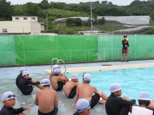 プールには水が溜まっており、拡声器を持った先生が話をしており、プールサイドには児童の皆さんが水泳帽をかぶり、水着姿で座って話を聞いている水泳指導のようすの写真