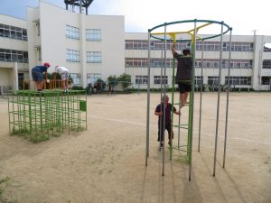 運動場に設置されている遊具のジャングルジムの上に2人の児童が登っており、手前の登り棒の遊具にも一人の児童が先生が見守る中、上まで登り、遊具の安全点検を行っている様子の写真