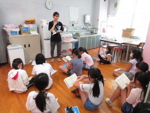 保健室に集まった女子児童が手に持った教科書を広げて座っており、子どもたちの前に立った養護教諭の先生が子どもたちに話をしている保健指導の様子の写真