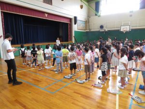 体育館で、子どもたちが手を後ろに組んで整列して並んでおり、舞台前に一人の先生が立って子どもたちに向けて話をしている終業式の様子の写真