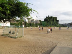 サッカーゴールのある運動場に地域の方々が集まって、両手を横に広げ、ラジオ体操をしている様子の写真