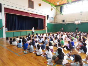 体育館で、子どもたちが整列して体育座りをしており、舞台前に一人の先生が立って子どもたちに向けて話をしている始業式の様子の写真