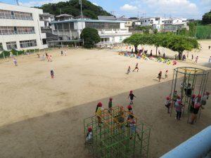 広々とした校庭で、走り回っている児童、ジャングルジムなどの遊具で遊んでいる児童、藤棚の下で遊んでいる児童などが写っている校庭の様子を写した写真