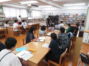 本棚に本が並んでいる図書室の机に参加者の方々が座って前方を見ており、前方のスペースで関係者の男性数名が刺股を持って、訓練を行っている様子を写した写真