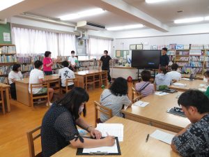 本棚に本が並んでいる図書室の机に参加者の方々が座っており、参加者の女性の一人が自席で立ち、発表しており、担当者の男性が話を聞いている研修の様子の写真