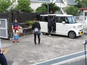 校門前の駐車スペースに白い軽自動車が止まっており、低学年の女子児童のお迎えに関係者の女性が来ている様子を写した写真