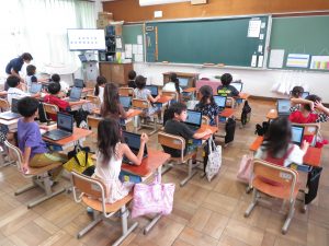 教室の自席についている子どもたちが自分の机の上にタブレットを広げており、前方の大型モニターに映像が写され、授業が行われている様子の写真