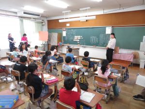 教室の黒板前に立って女性の先生が授業をする中、子どもたちが自席について授業を受けており、窓側には書類を手に持ち、授業を見ている先生方が写っている校内研究授業の様子の写真