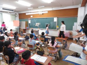 教室の黒板前に立って女性の先生が授業をする中、黒板前に立って発表をしている女子児童、自席について授業を受けているクラスメート、左側にはメモを取りながら授業を参観している先生方がずらりと並んでいる校内研究授業の様子の写真