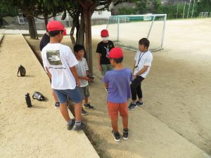 運動場の木陰に児童5名と先生が集まり、話し合いをしている様子の写真
